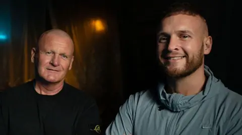 BBC/Double Act Productions Andrew and Macaulay Dodd pictured standing next to each other. Both looks at the camera and smiles. Andrew wears a black top and Macaulay a light blue sports zip-up top. It is a head and shoulders shot of them.