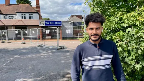 Wael, a man with curly hair, is wearing a navy blue jumper. He stands in front of fences outside the Bell hotel in Epping which has temporary metal fences outside.