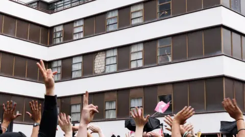Dan Kitwood/Getty Images Anti Fascist protesters cheers and wave as men believed to be migrants gesture from a window of the Barbican Thistle Hotel as anti-immigration, 'stand up to racism' and 'anti-fascist' groups all gather outside on August 2, 2025 in London, England.