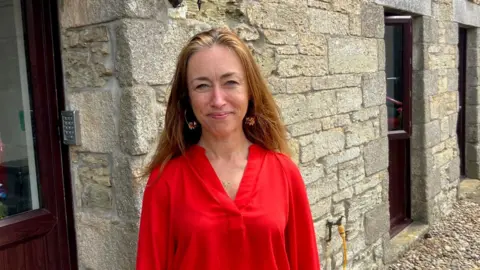 A woman stands in front of a granite building smiling. She has long auburn hair and is wearing a red blouse.