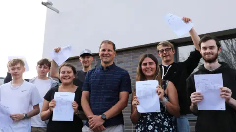 Deep South Media A group of Arnewood School students stand outside the school building with Mark Colman, head of sixth form. Everyone is smiling at the camera. There are three male students and one female student to the left of Mr Colman, holding pieces of paper with their results on either in front of their chest or in the air. Another two male students and a female student stand to the right of Mr Colman holding their results. Mr Colman has short, light brown hair, wears a short sleeved dark blue striped shirt and beige chino trousers. He has his hands crossed in front of him in a relaxed pose. 