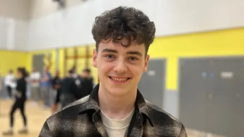 A young man with curly brown hair which is cut short at the sides and longer on top. He is smiling at the camera and wearing a brown and tan coloured flannel shirt. Behind him, out of focus, is a school hall with other pupils milling around.