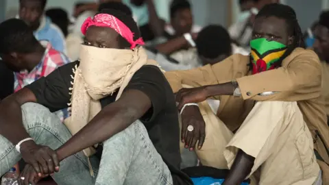 Francesco Tosto/BBC Migrant arrivals from North Africa sit wearing masks in a crowded hall in Crete