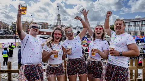 Julian Preece Photography Five people, two men and three women, cheer and raise their arms in the air. They are all wearing the same outfit of multicoloured polka dot plastic skirts over dark shorts and white T shirts with small coloured dots on them. They are all holding what looks like pints of beer and behind them Bristol's harbourside ampitheatre is visible