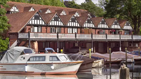 Historic England Archive The boathouse, with rows of gable roofs, a balcony stretching across the front of the building with cars parked underneath. In the foreground are modern boats moored on the river.