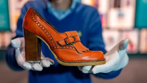 Plaster Communications An elegant high-heeled shoe is held in the hands of a member of staff at the Clarks museum in Somerset. He is wearing white gloves. The shoe has a thick heel and intricate stitching and is light brown in colour