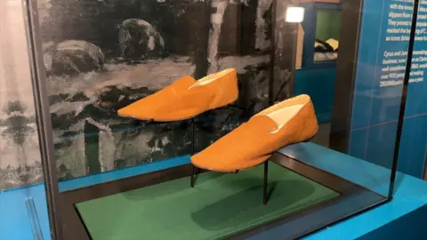 Picture of a pair of light brown slippers made from a thin material in a glass case at a museum. They are each on a small metal pole holding them in place, above a rectangular piece of green material