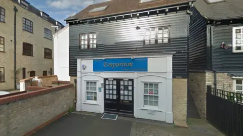 Google The entrance to a shop named 'Emporium', featuring a black door with glass panes and a small accessibility ramp. The building has a white brick lower facade and a black wooden upper facade, with windows on both levels.
