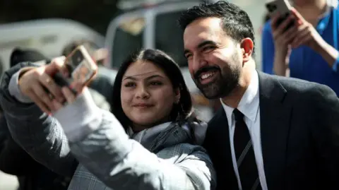 Reuters Zohran Mamdani poses for selfie with young New Yorker. He is wearing a dark suit, white shirt and dark tie with silver diagonal stripes. The woman is in her teens and she is holding the camera as they both smile at the lens