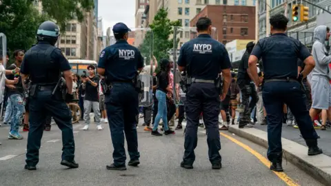 Reuters Four police officers have their backs to us as they provide security in Union Square as popular live streamer, not shown, stages a giveaway. They have NYPD on their shirts and one of them wears a helmet. 
