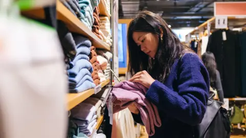 Getty Images A woman with loose long dark hair in a dark blue jumper is looking at clothes she has taken from a shop shelf